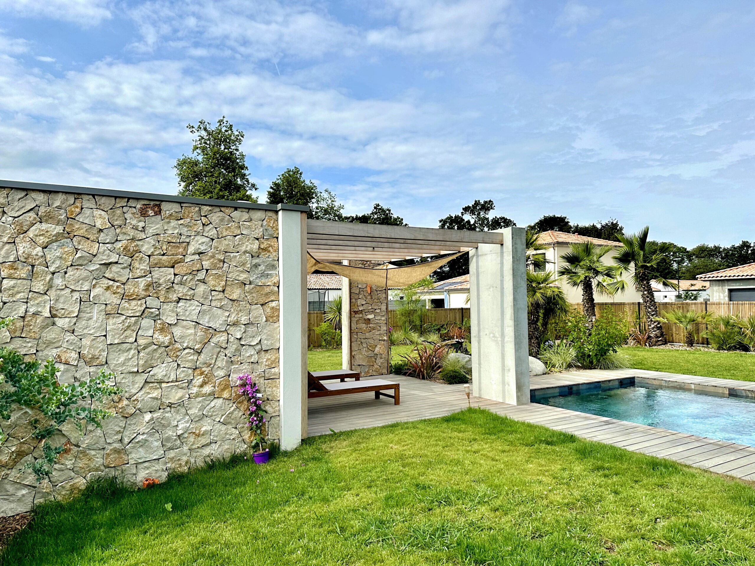Jardin Eden tropical à Challans avec parement naturel mur en pierre de taille, plage de piscine en bois, massifs style tropical