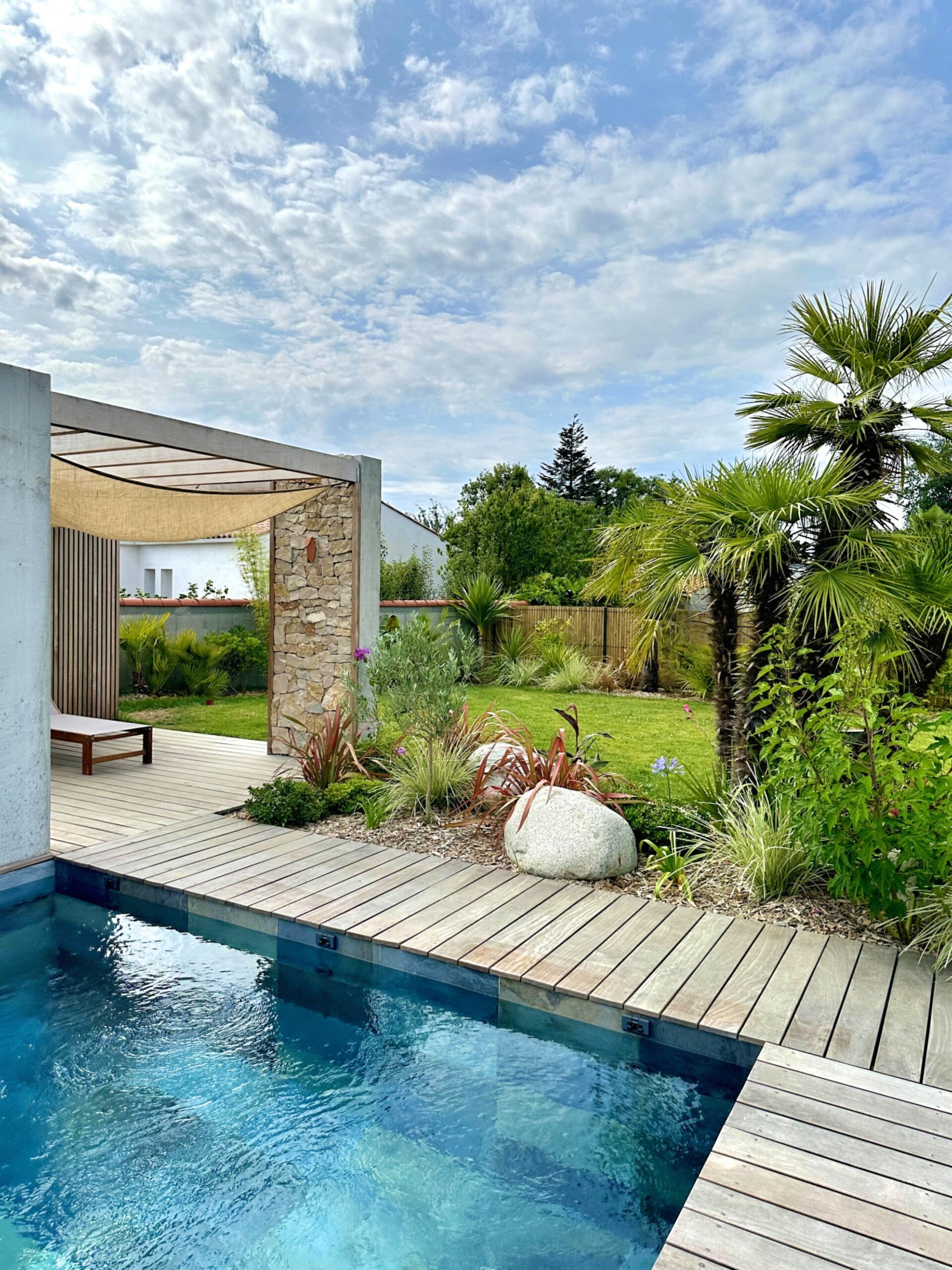Jardin Eden tropical à Challans avec mur en pierre de taille, plage de piscine en bois, massifs style tropical avec roches des seychelles