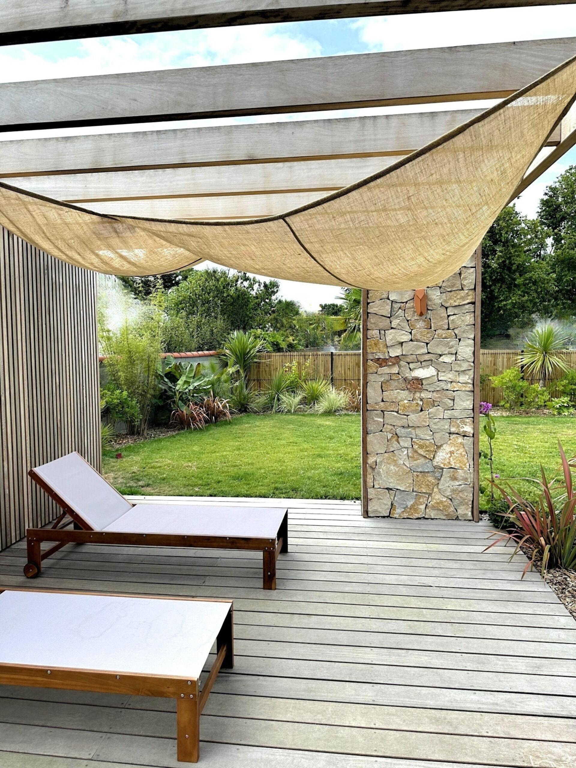 Jardin Eden tropical à Challans avec mur en pierre de taille, plage de piscine en bois, massifs, palmiers style tropical avec roches des seychelles
